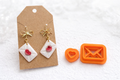 Envelope and heart polymer clay cutters sit on a white background next to a set of completed polymer clay envelope earrings sealed with a red heart on bow earring hooks. 